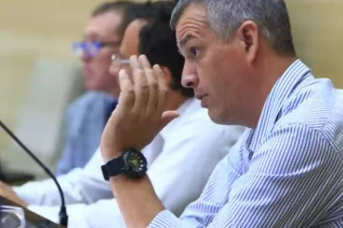 Hombre de cabello corto y canoso con camisa de rayas sentado en una mesa junto a otras personas en lo que parece ser una reunión o conferencia