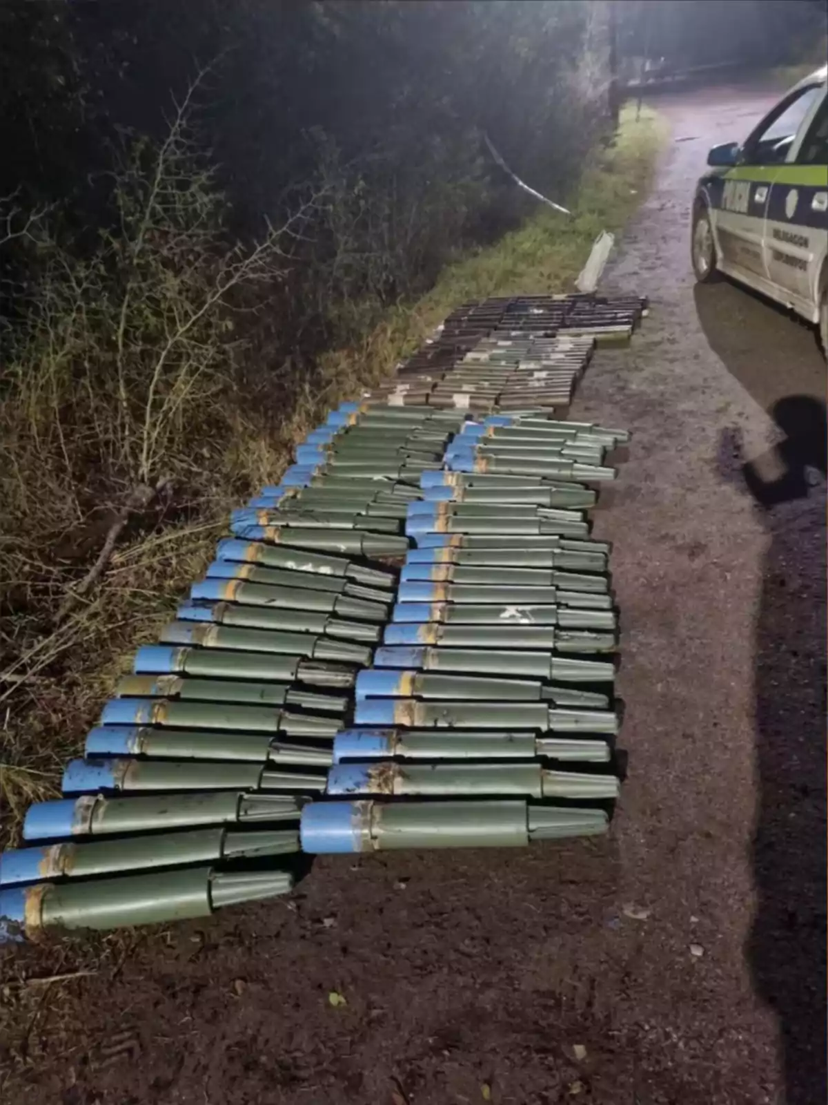 Varios proyectiles de color verde con punta azul están alineados sobre el suelo junto a un camino y un vehículo policial