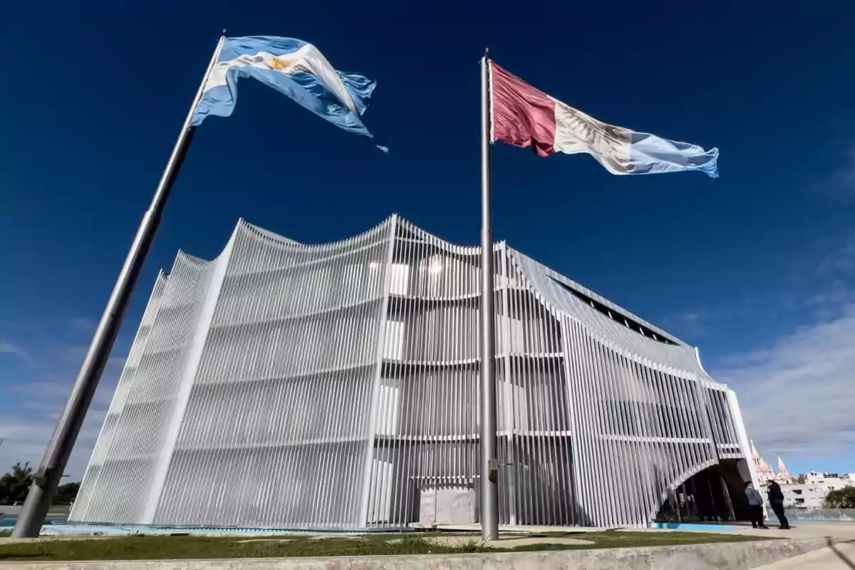 Edificio moderno con fachada de lamas verticales blancas y dos banderas ondeando al frente bajo un cielo azul