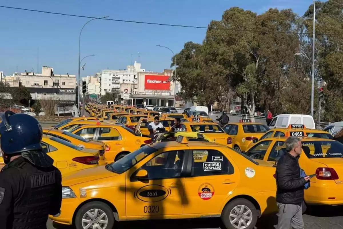 Numerosos taxis amarillos están detenidos en una calle de la ciudad mientras varias personas y un policía observan la escena Numerosos taxis amarillos están detenidos en una calle de la ciudad mientras varias personas y un policía observan la escena