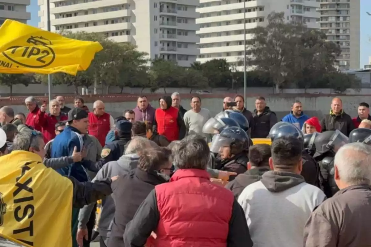 A group of people gathers on the street in front of several tall buildings; some are wearing colorful vests and jackets, there are police officers with helmets, and a yellow flag is waving on the left. A group of people gathers on the street in front of several tall buildings; some are wearing colorful vests and jackets, there are police officers with helmets, and a yellow flag is waving on the left.
