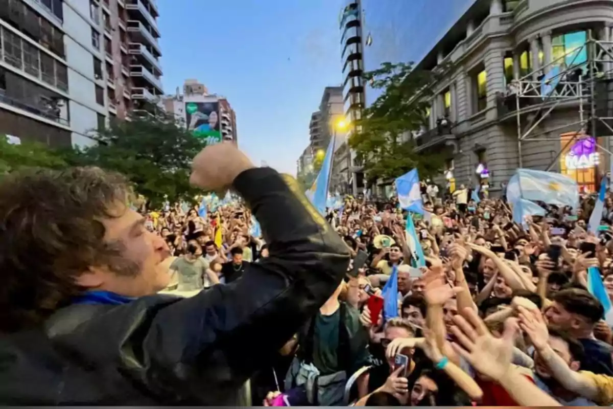 A man in a leather jacket raises his fist in front of a cheering crowd on a street surrounded by buildings while several people wave Argentine flags. A man in a leather jacket raises his fist in front of a cheering crowd on a street surrounded by buildings while several people wave Argentine flags.
