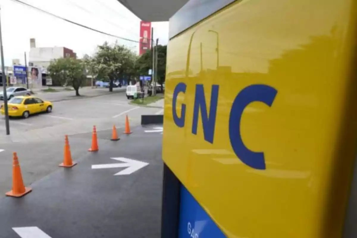 Cartel amarillo de GNC en una estación de servicio con conos naranjas y un taxi amarillo en la calle