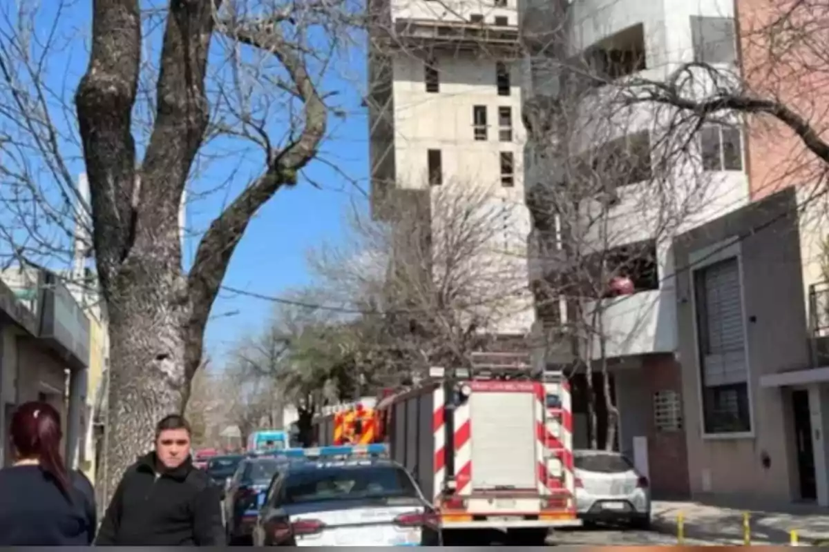 Camión de bomberos y patrullas estacionados en una calle arbolada frente a un edificio, con personas observando la escena Camión de bomberos y patrullas estacionados en una calle arbolada frente a un edificio, con personas observando la escena