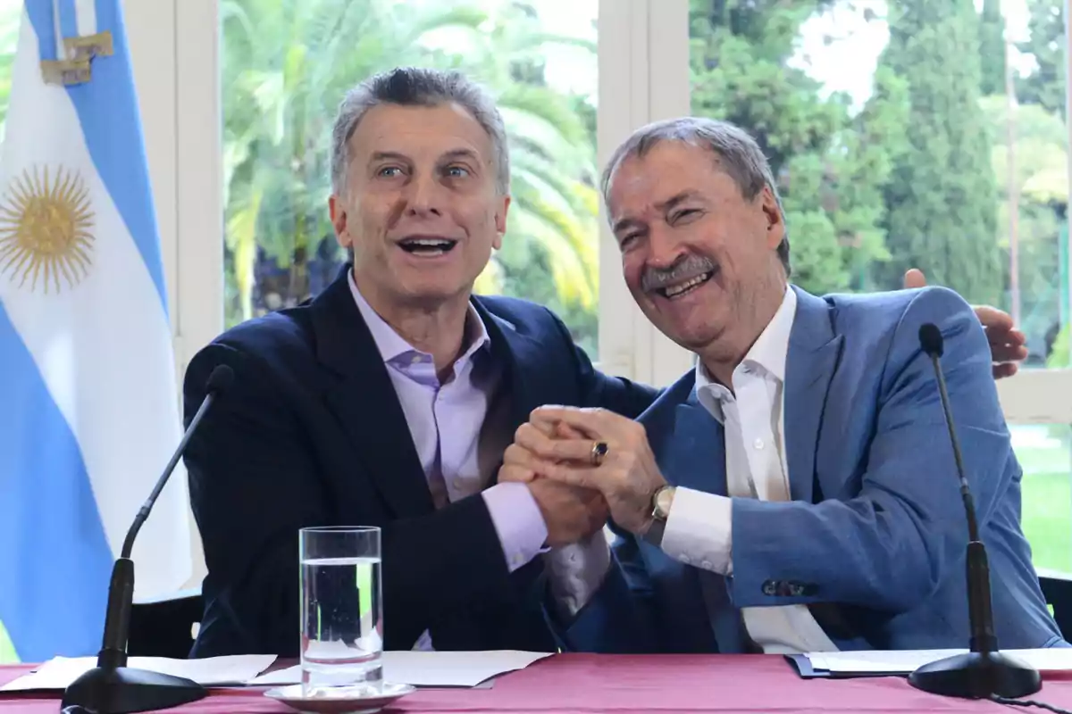 Dos hombres sonríen y se saludan efusivamente sentados en una mesa con micrófonos y un vaso de agua, al fondo se ve una bandera argentina y vegetación exterior Dos hombres sonríen y se saludan efusivamente sentados en una mesa con micrófonos y un vaso de agua, al fondo se ve una bandera argentina y vegetación exterior