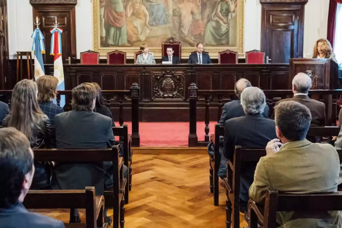 People seated in a courtroom watching three judges on the main bench with Argentine flags to the side and a woman speaking from a podium