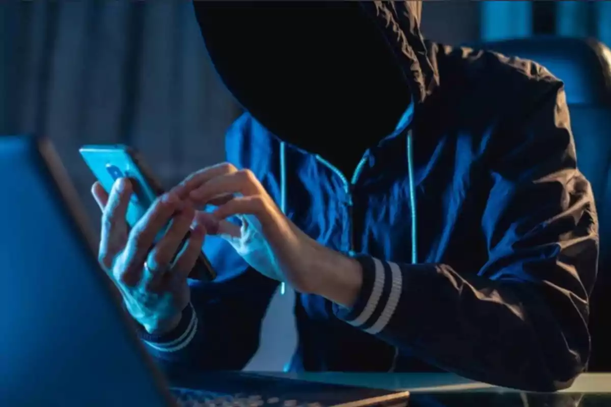 Person wearing a hoodie using a mobile phone in front of a computer in a dark environment