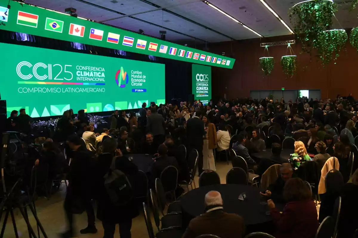 Auditorium filled with people sitting and standing during an international climate conference, with a large green screen in the background displaying the event name and several flags from different countries at the top. Auditorium filled with people sitting and standing during an international climate conference, with a large green screen in the background displaying the event name and several flags from different countries at the top.