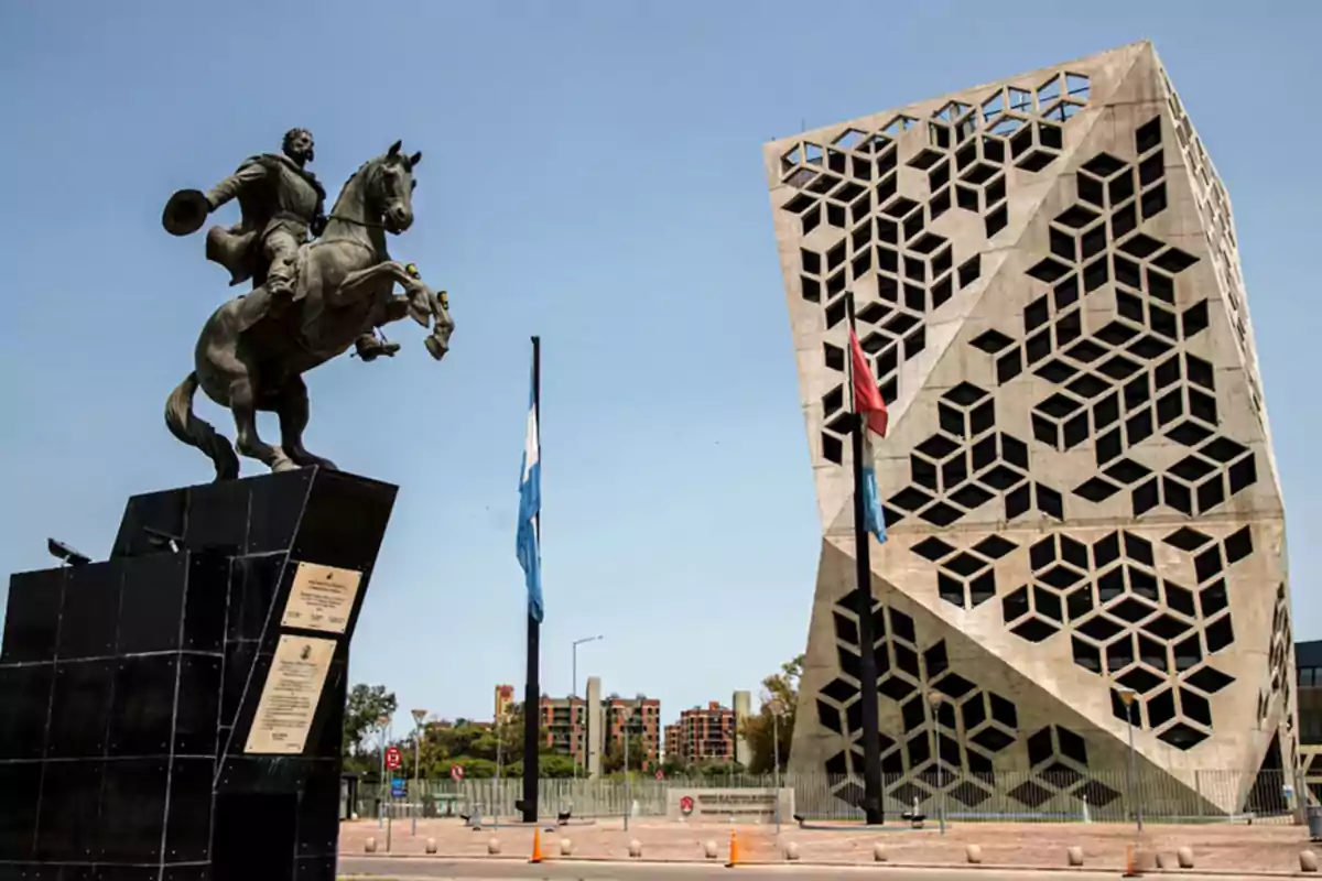 Escultura ecuestre frente a un edificio moderno con diseño geométrico y banderas ondeando en un día soleado