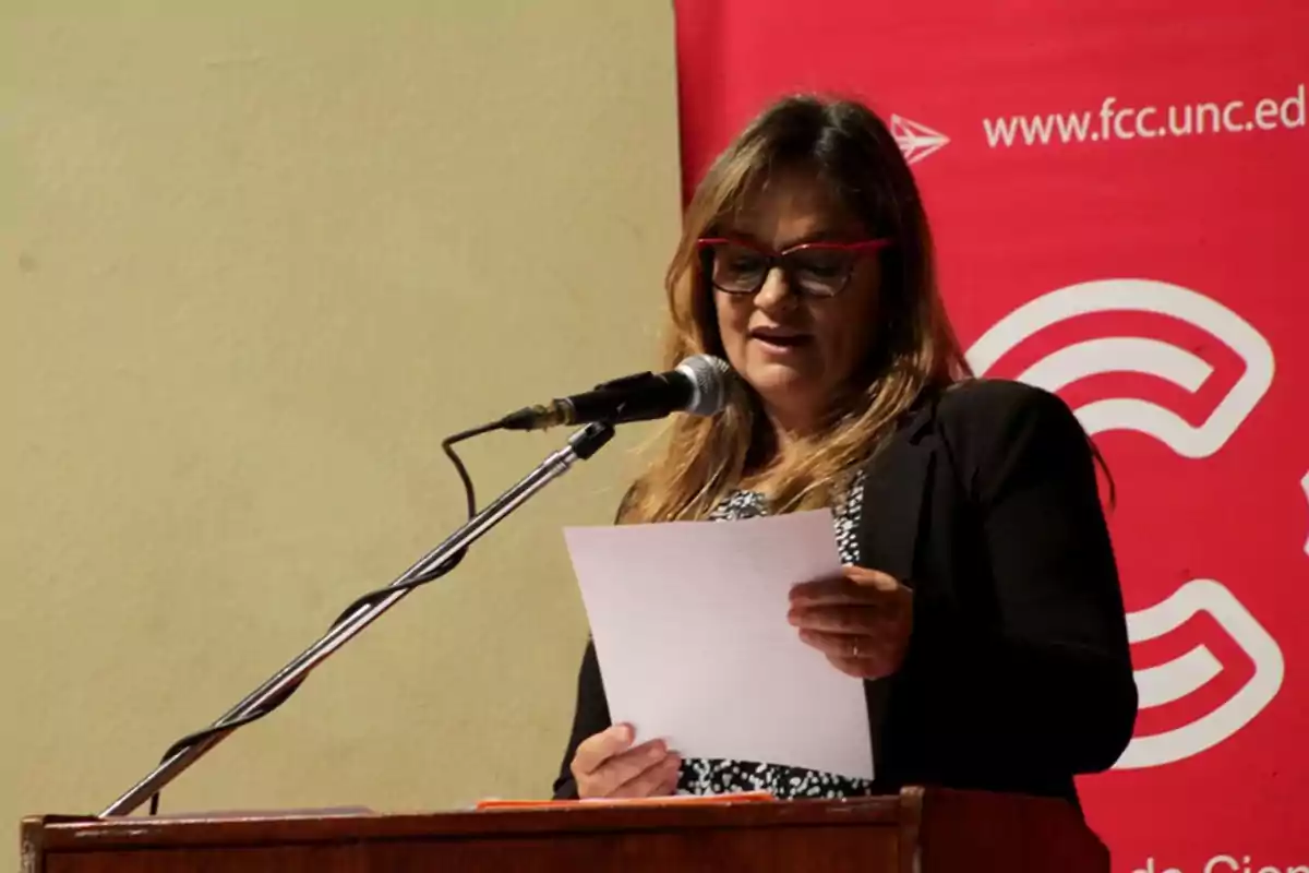 Mujer con gafas rojas leyendo un documento frente a un micrófono en un podio con fondo rojo de una universidad Mujer con gafas rojas leyendo un documento frente a un micrófono en un podio con fondo rojo de una universidad