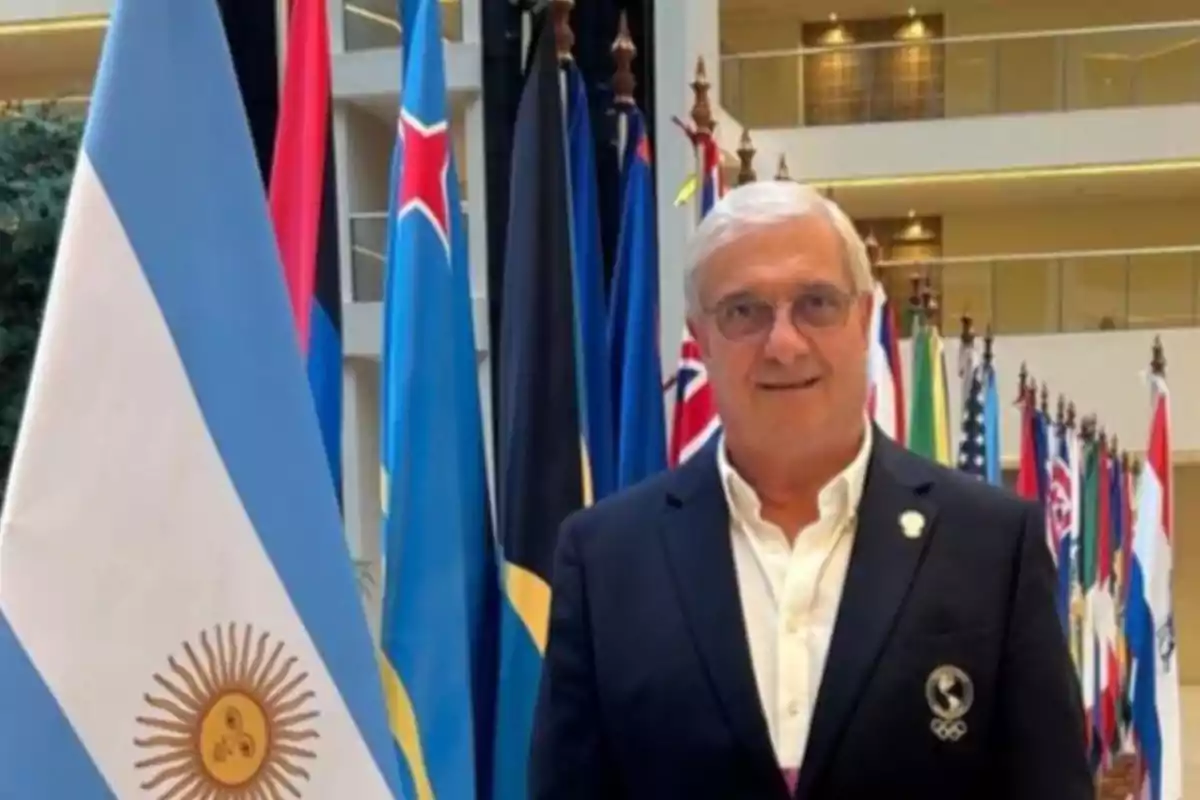 Hombre de cabello canoso y gafas posando junto a la bandera de Argentina y varias banderas de diferentes países en un edificio moderno Hombre de cabello canoso y gafas posando junto a la bandera de Argentina y varias banderas de diferentes países en un edificio moderno
