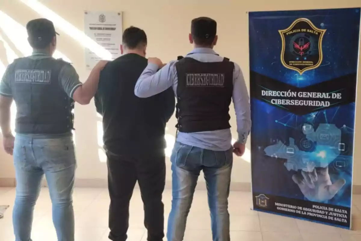 Dos agentes de ciberseguridad escoltan a una persona mientras posan de espaldas junto a un cartel de la Dirección General de Ciberseguridad de la Policía de Salta Dos agentes de ciberseguridad escoltan a una persona mientras posan de espaldas junto a un cartel de la Dirección General de Ciberseguridad de la Policía de Salta