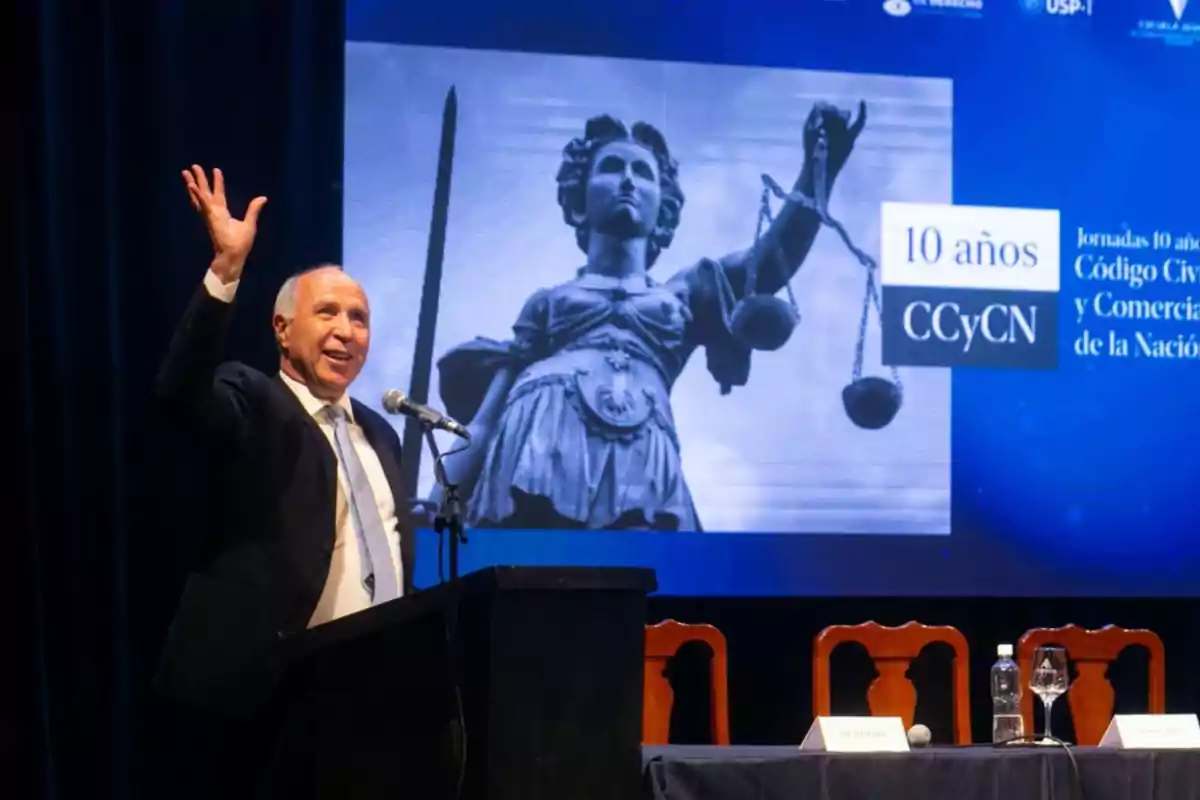 Man in a suit speaking at a podium and waving with his hand raised during an event, with an image of the statue of justice and a sign that reads Man in a suit speaking at a podium and waving with his hand raised during an event, with an image of the statue of justice and a sign that reads