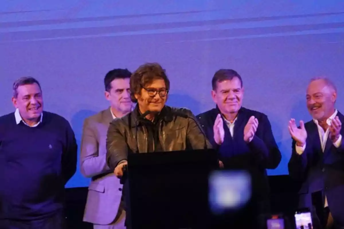 Cinco hombres de pie en un escenario, uno de ellos hablando en un podio mientras los otros aplauden y sonríen, con fondo azul.