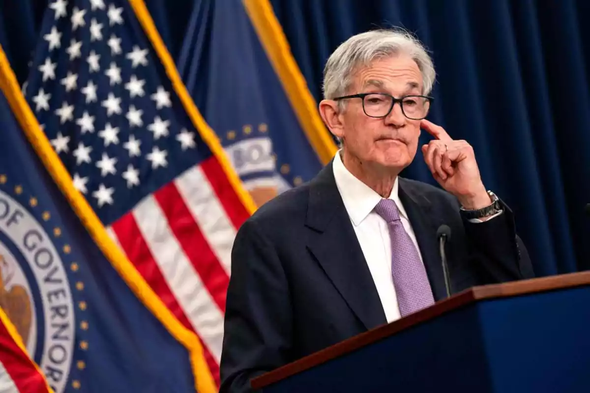 Hombre de cabello canoso y gafas hablando en un podio con banderas de Estados Unidos al fondo