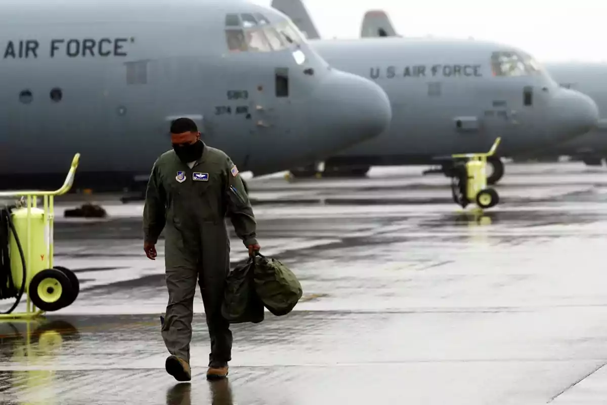 Un militar camina por la pista de un aeropuerto bajo la lluvia llevando una bolsa mientras aviones de la Fuerza Aérea de Estados Unidos están estacionados al fondo.