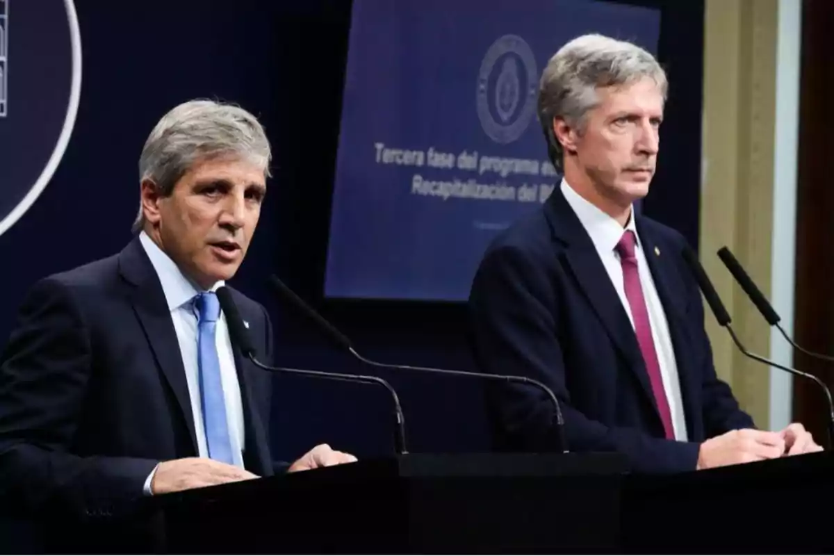 Two men in suits speaking at a podium during a press conference with a blue background and an institutional sign. Two men in suits speaking at a podium during a press conference with a blue background and an institutional sign.