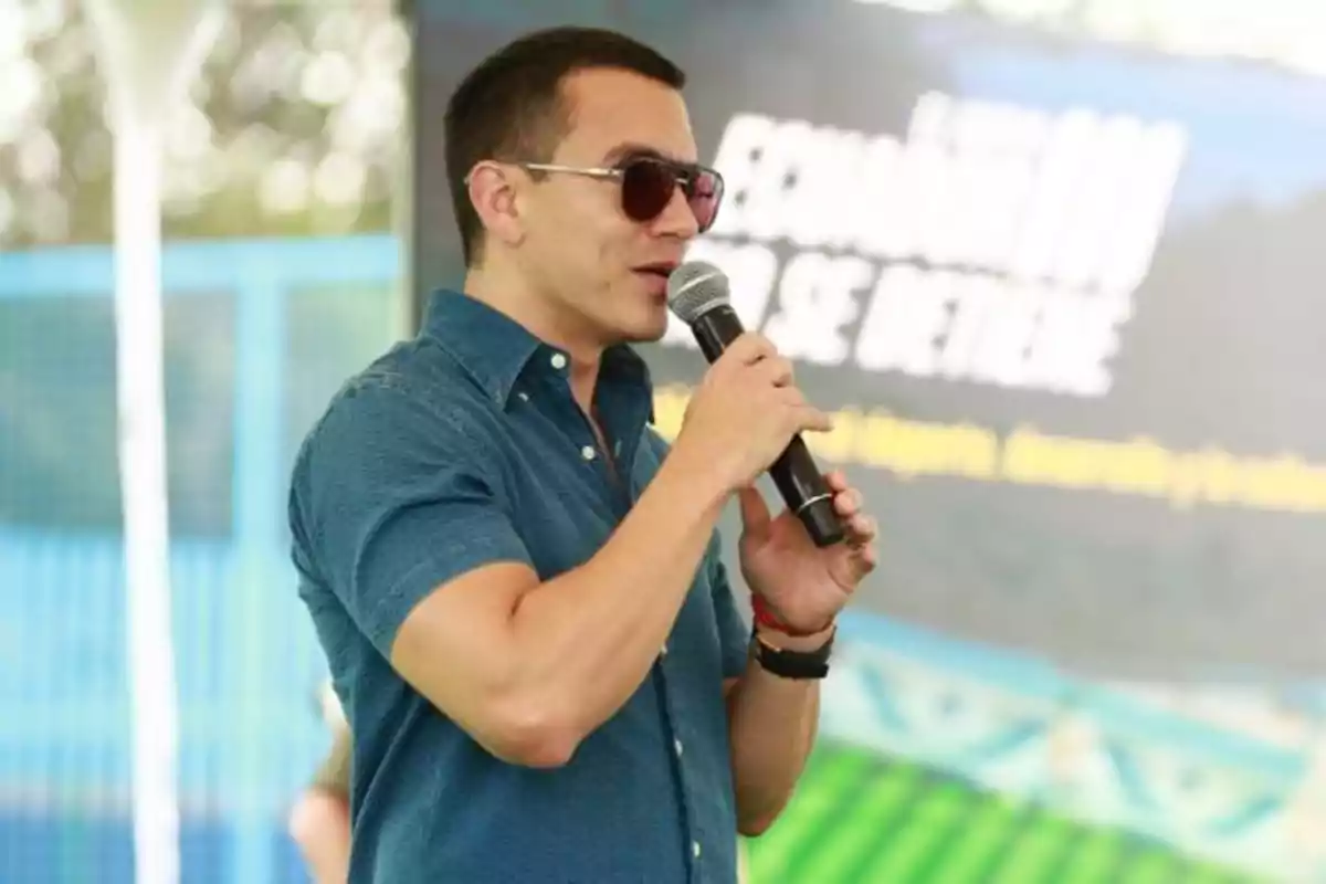 Hombre con gafas de sol y camisa azul hablando por micrófono en un evento al aire libre