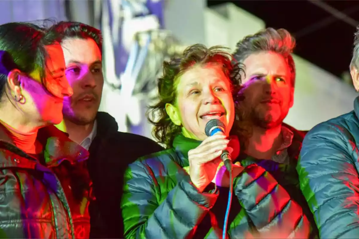 Group of people in warm jackets under colored lights, a woman in the center is holding a microphone and appears to be speaking or singing. Group of people in warm jackets under colored lights, a woman in the center is holding a microphone and appears to be speaking or singing.