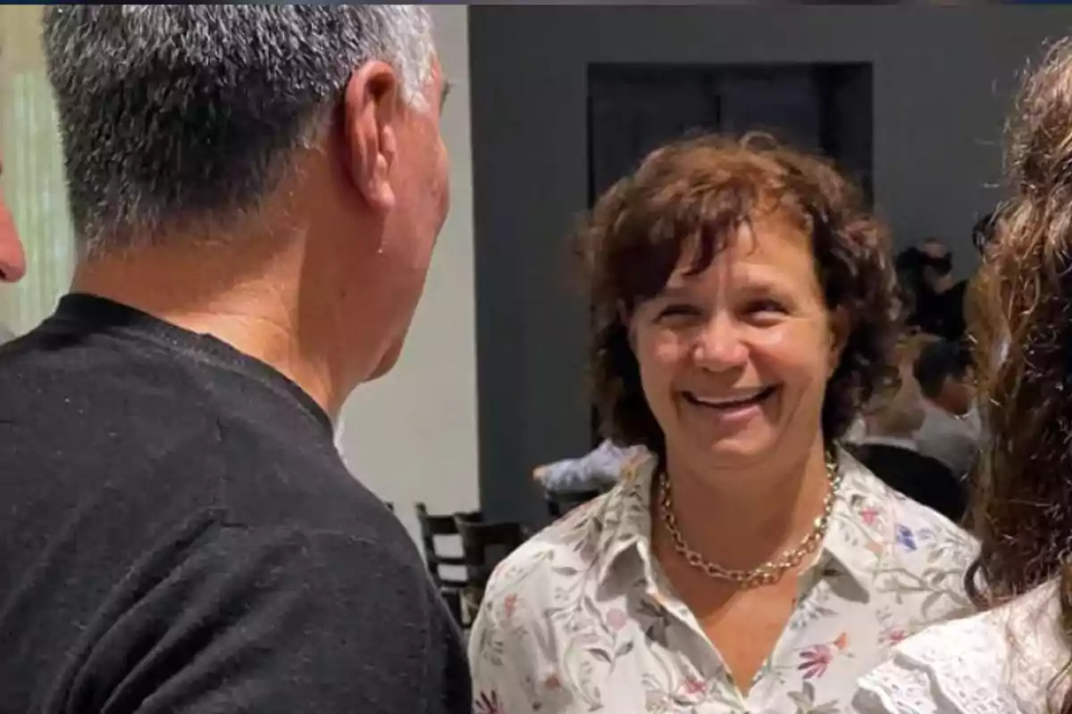 Smiling woman in a floral jersey and gold necklace talking with other people in an indoor setting Smiling woman in a floral jersey and gold necklace talking with other people in an indoor setting