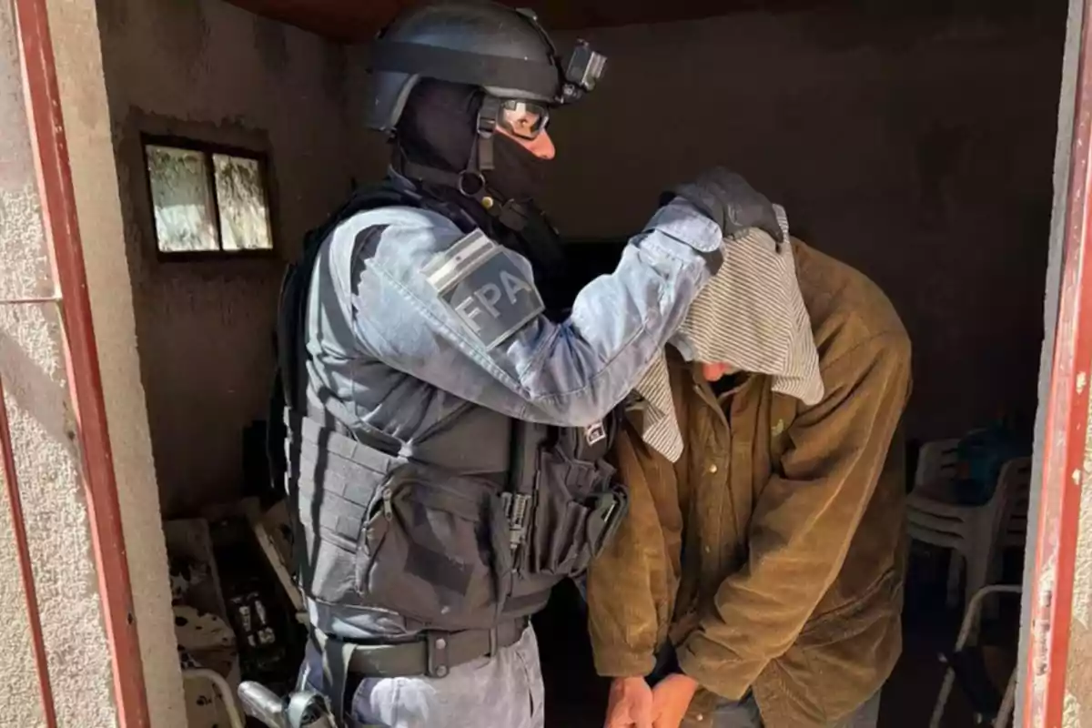 A police officer in tactical uniform detains a person with handcuffed hands and a cloth covering their head inside a room. A police officer in tactical uniform detains a person with handcuffed hands and a cloth covering their head inside a room.
