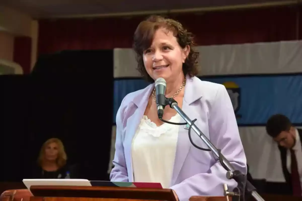 Short, curly-haired woman wearing a lilac blazer speaking into a microphone at a podium