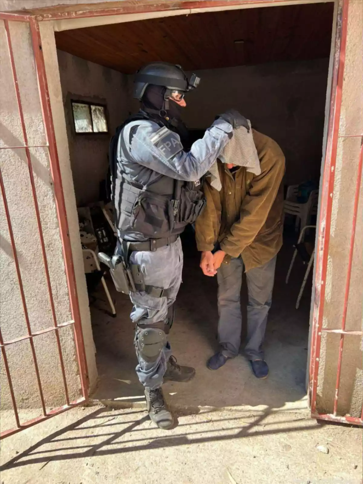 A police officer in tactical gear detains a handcuffed person and covers their head with a cloth at the entrance of a house. A police officer in tactical gear detains a handcuffed person and covers their head with a cloth at the entrance of a house.