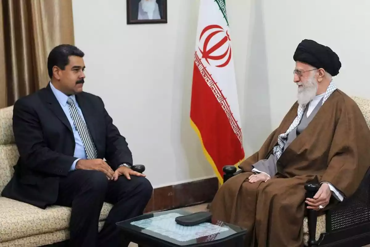 Two men sit talking in a room next to an Iranian flag.