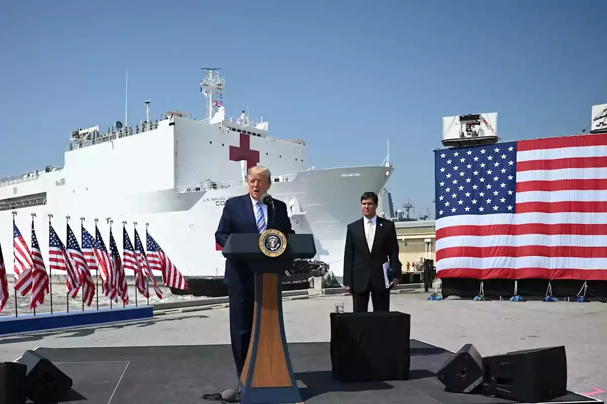 Un hombre de traje habla en un podio presidencial frente a un barco hospital de la marina estadounidense, acompañado de varias banderas de Estados Unidos y una gran bandera al fondo