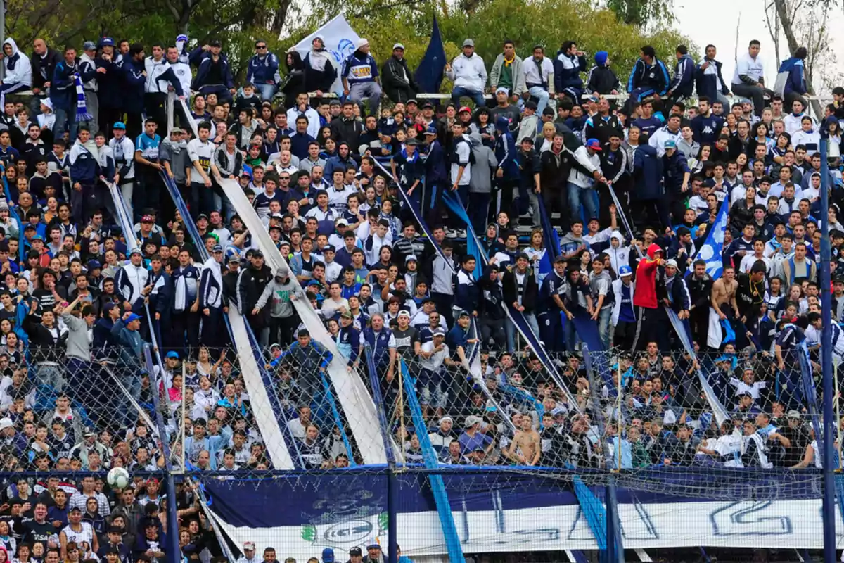 Aficionados de un equipo de fútbol llenan las gradas del estadio, vestidos con ropa en tonos azul y blanco, animando y mostrando banderas y pancartas.