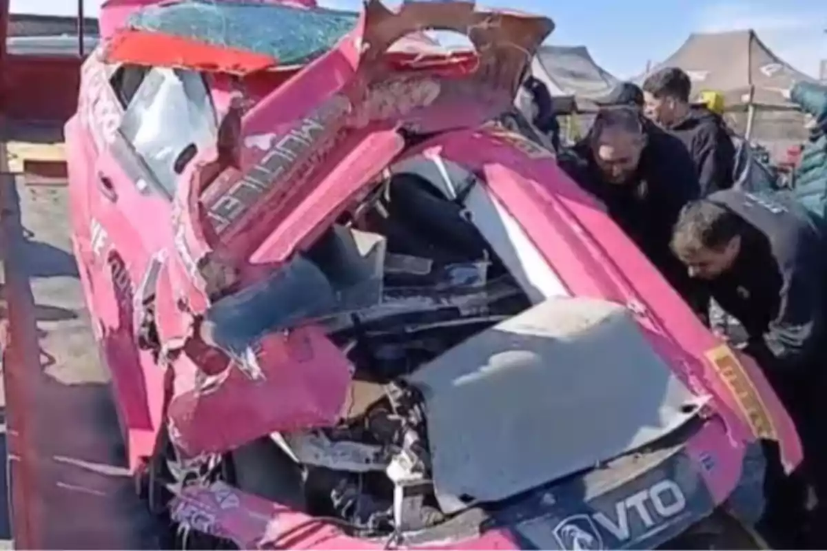 Auto de carreras color rosa completamente destrozado mientras varias personas lo observan y revisan en un área de boxes o taller al aire libre Auto de carreras color rosa completamente destrozado mientras varias personas lo observan y revisan en un área de boxes o taller al aire libre