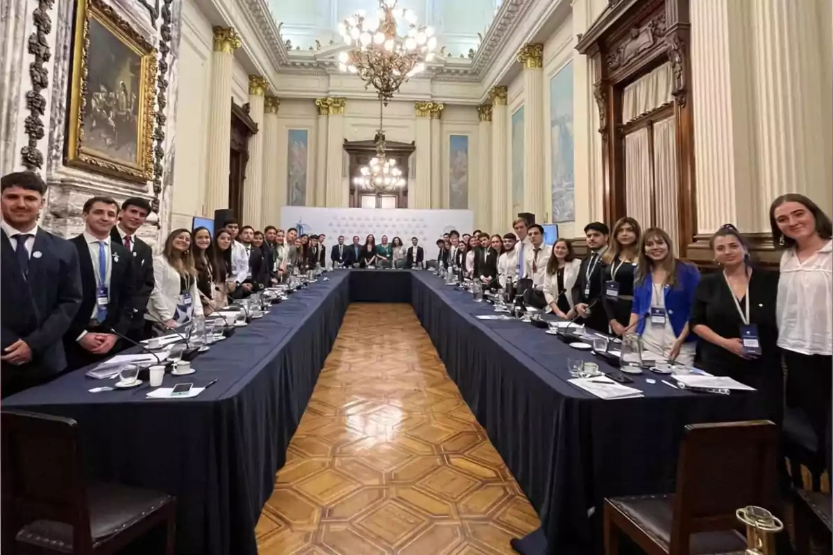 Un grupo numeroso de personas vestidas de manera formal está de pie alrededor de una mesa en forma de U en un elegante salón con techos altos, lámparas de araña y detalles arquitectónicos clásicos Un grupo numeroso de personas vestidas de manera formal está de pie alrededor de una mesa en forma de U en un elegante salón con techos altos, lámparas de araña y detalles arquitectónicos clásicos