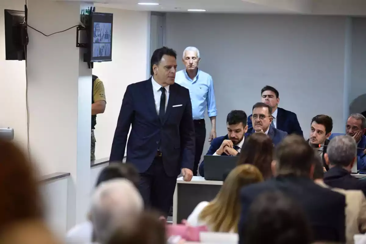 A man in a dark suit walks through a room full of seated people, some of whom are watching alertly while others are focused on their computers and papers, in an environment that appears to be a hearing or trial. A man in a dark suit walks through a room full of seated people, some of whom are watching alertly while others are focused on their computers and papers, in an environment that appears to be a hearing or trial.