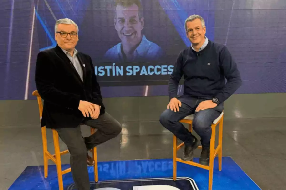 Two men sitting on tall stools in a television studio with a background screen displaying the name Agustín Spaccesi and an image of a smiling person. Two men sitting on tall stools in a television studio with a background screen displaying the name Agustín Spaccesi and an image of a smiling person.
