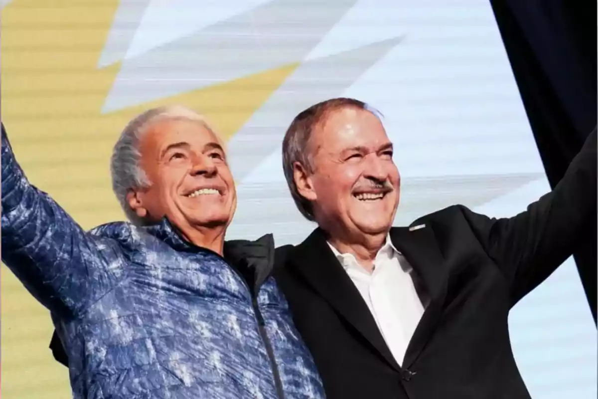 Dos hombres mayores sonríen y levantan los brazos en señal de celebración frente a un fondo claro con formas geométricas. Dos hombres mayores sonríen y levantan los brazos en señal de celebración frente a un fondo claro con formas geométricas.