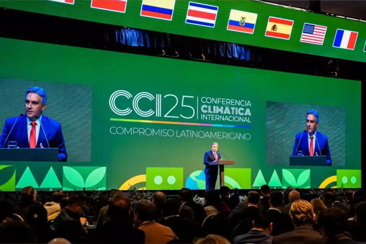A man in a blue suit and red tie gives a speech on stage during an international climate conference, with a large green screen in the background displaying flags of various countries and the text A man in a blue suit and red tie gives a speech on stage during an international climate conference, with a large green screen in the background displaying flags of various countries and the text