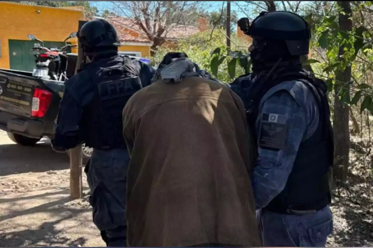 Three heavily armed police officers escort a person wearing a brown jacket and with their head covered, as they walk next to a pickup truck in a residential area. Three heavily armed police officers escort a person wearing a brown jacket and with their head covered, as they walk next to a pickup truck in a residential area.