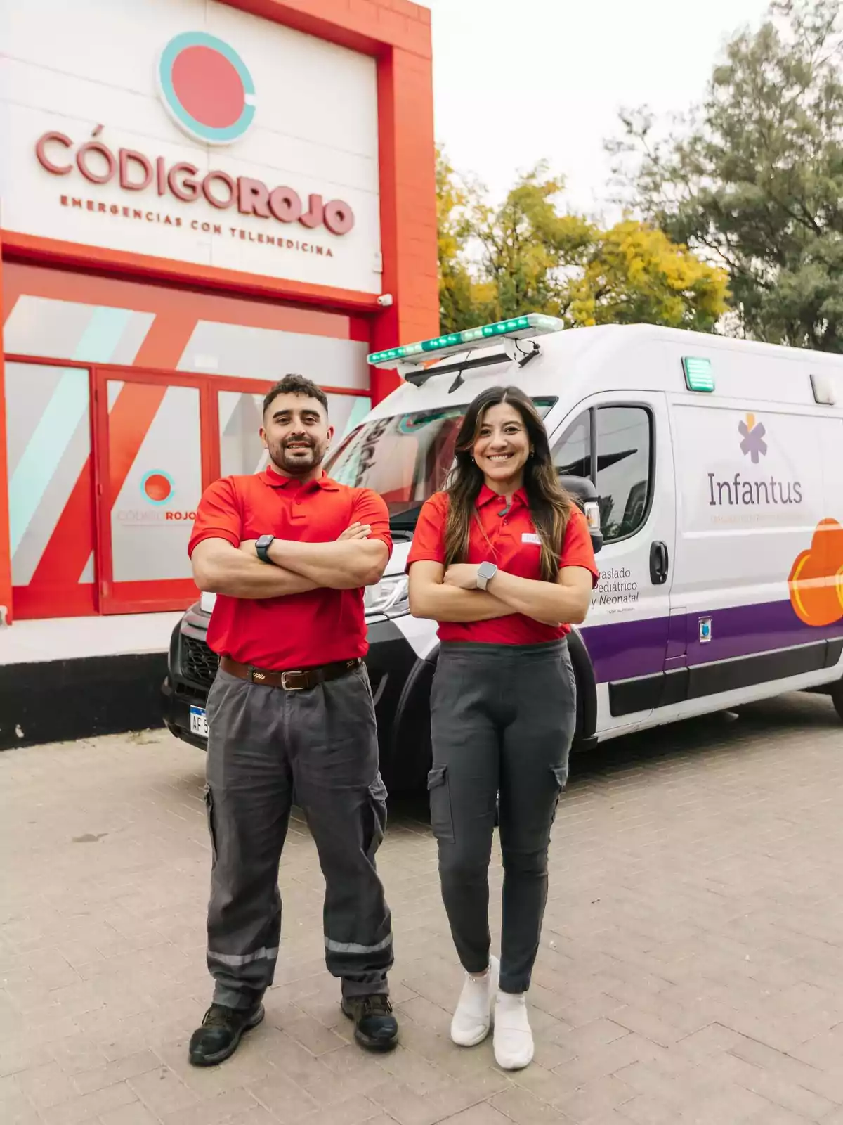 Two paramedics in red uniforms and gray pants pose smiling with their arms crossed in front of an ambulance and an emergency building called Código Rojo. Two paramedics in red uniforms and gray pants pose smiling with their arms crossed in front of an ambulance and an emergency building called Código Rojo.