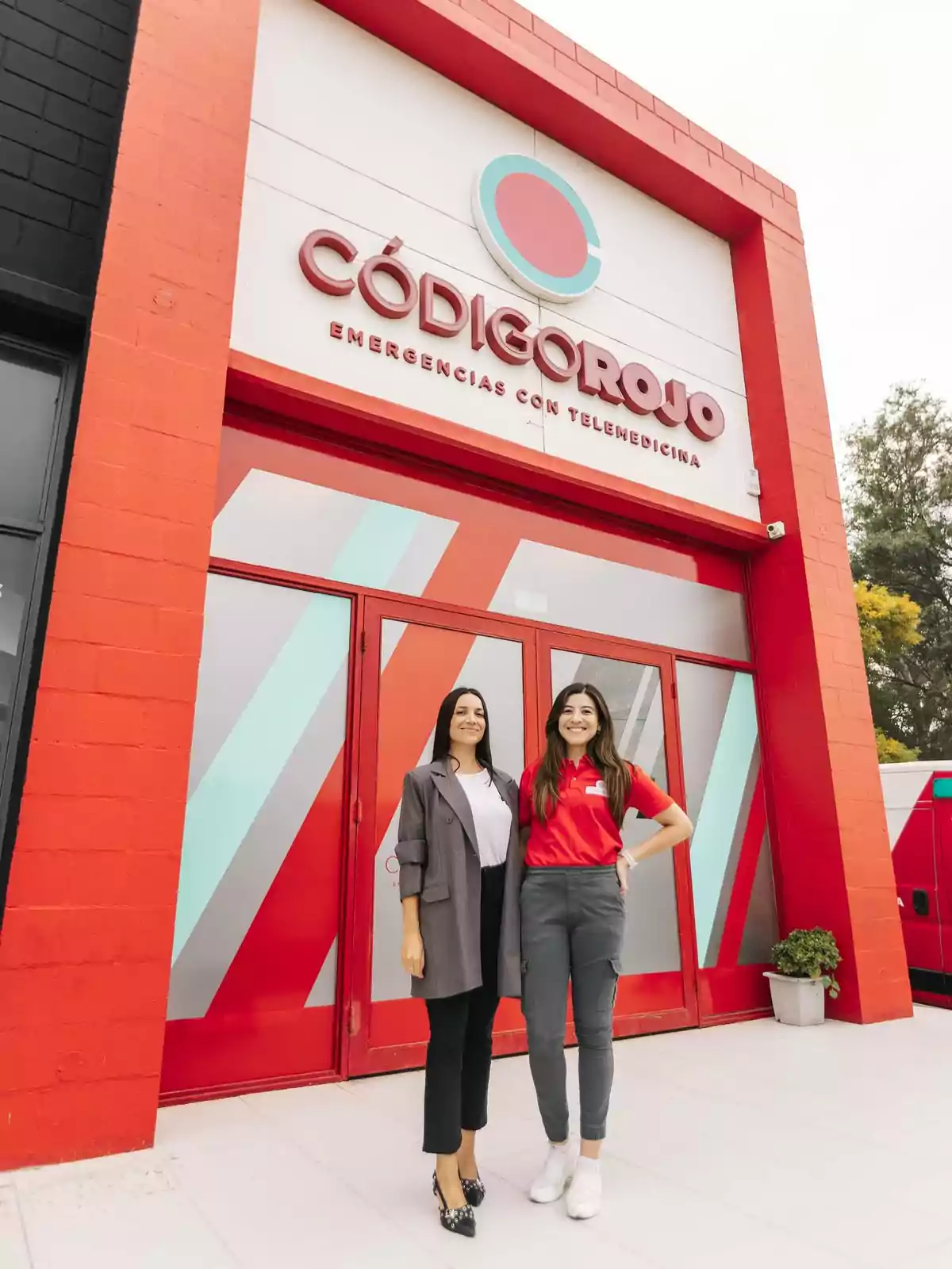 Two smiling women pose in front of the entrance to an emergency clinic called Code Red, featuring a modern facade in red tones and geometric details. Two smiling women pose in front of the entrance to an emergency clinic called Code Red, featuring a modern facade in red tones and geometric details.