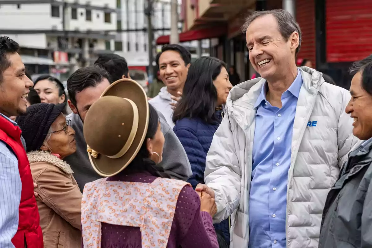 Un hombre sonriente con chaqueta clara saluda a una mujer con sombrero marrón mientras varias personas los rodean y observan en una calle urbana