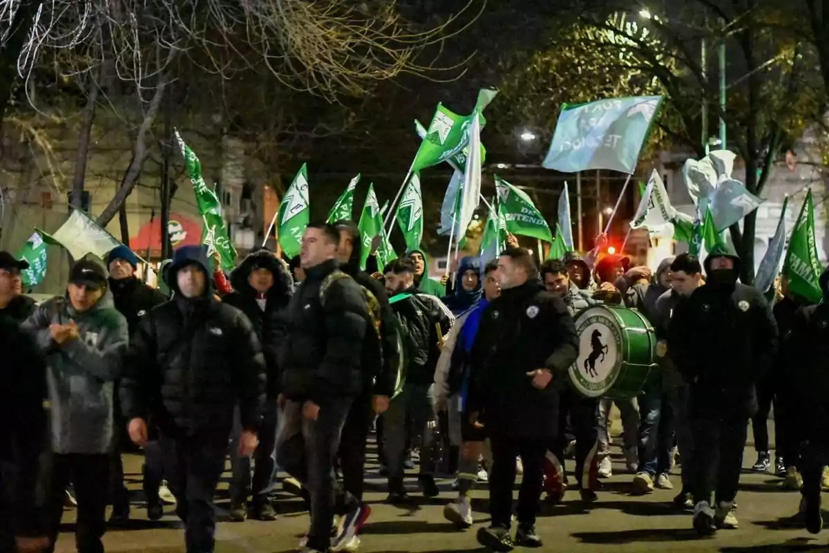 A group of people walks down the street at night with green flags and a bass drum, all bundled up in jackets and hats. A group of people walks down the street at night with green flags and a bass drum, all bundled up in jackets and hats.