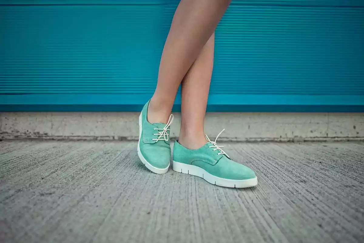 Person standing with crossed legs wearing green shoes with white soles on a concrete floor in front of a blue wall Person standing with crossed legs wearing green shoes with white soles on a concrete floor in front of a blue wall