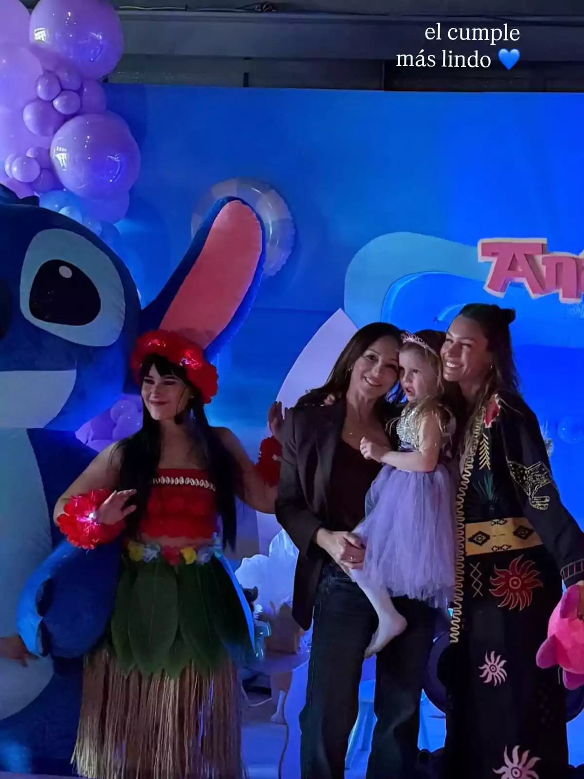 Grupo de personas celebrando una fiesta infantil con decoración de globos morados y un personaje disfrazado de Stitch, una niña con vestido lila y adultos sonrientes posando juntos Grupo de personas celebrando una fiesta infantil con decoración de globos morados y un personaje disfrazado de Stitch, una niña con vestido lila y adultos sonrientes posando juntos