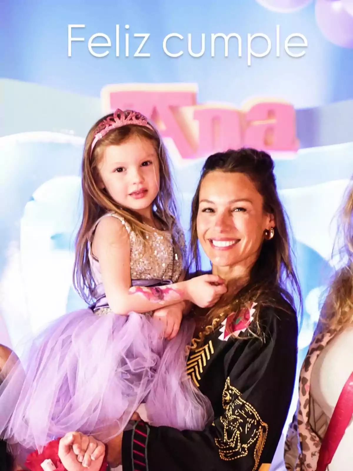 Niña con vestido y corona lila siendo cargada por una mujer sonriente en una fiesta de cumpleaños con decoración colorida y el texto feliz cumple al fondo Niña con vestido y corona lila siendo cargada por una mujer sonriente en una fiesta de cumpleaños con decoración colorida y el texto feliz cumple al fondo