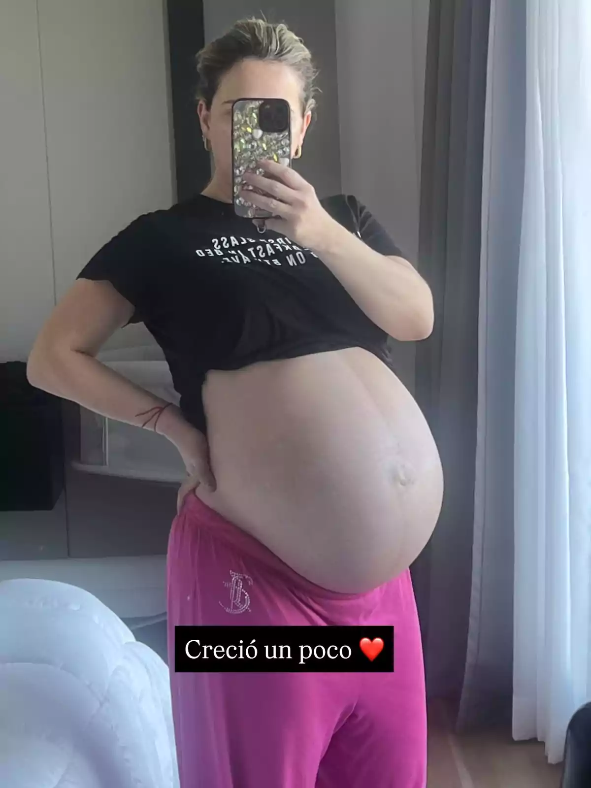 Mujer embarazada tomándose una selfie frente al espejo, usando pantalones rosas y camiseta negra, con la frase Creció un poco y un emoji de corazón rojo