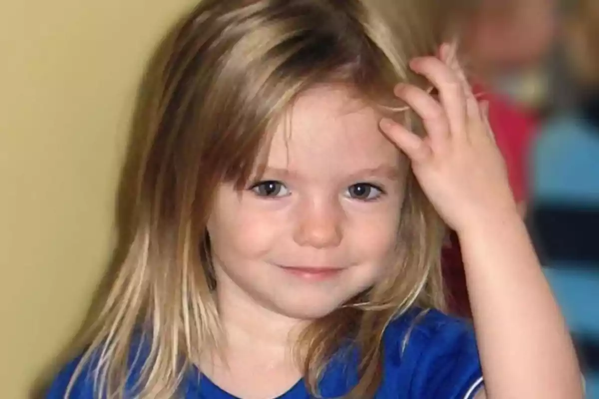 Niña pequeña de cabello rubio y ojos claros sonriendo y tocándose la cabeza con una mano