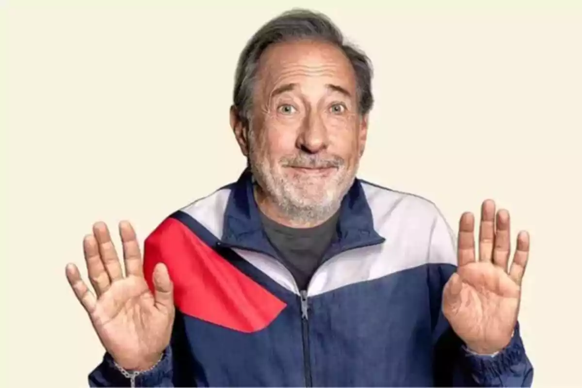 Hombre mayor con barba y cabello canoso sonríe y levanta ambas manos con las palmas hacia adelante viste una chaqueta deportiva azul con rojo y blanco sobre un fondo claro Hombre mayor con barba y cabello canoso sonríe y levanta ambas manos con las palmas hacia adelante viste una chaqueta deportiva azul con rojo y blanco sobre un fondo claro