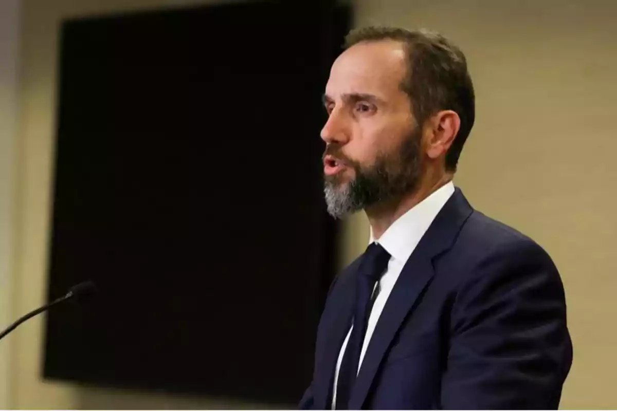 Hombre de barba y traje oscuro hablando frente a un micrófono en un entorno formal Hombre de barba y traje oscuro hablando frente a un micrófono en un entorno formal