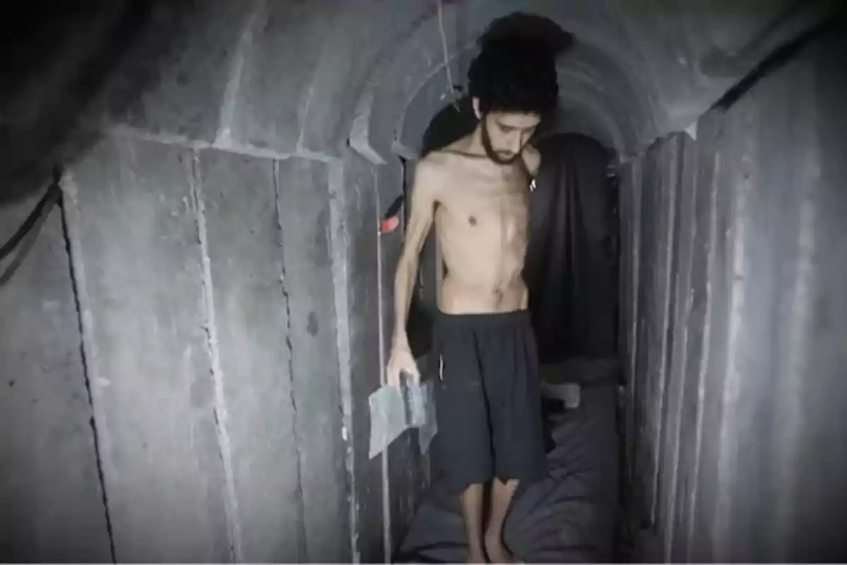 Hombre delgado con el torso descubierto y pantalones cortos oscuros dentro de un túnel de concreto sosteniendo una botella de agua