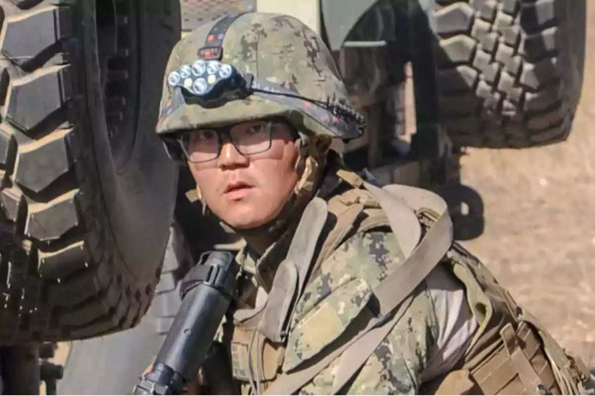 Soldier in camouflage uniform and helmet with flashlight, holding a weapon next to a military vehicle
